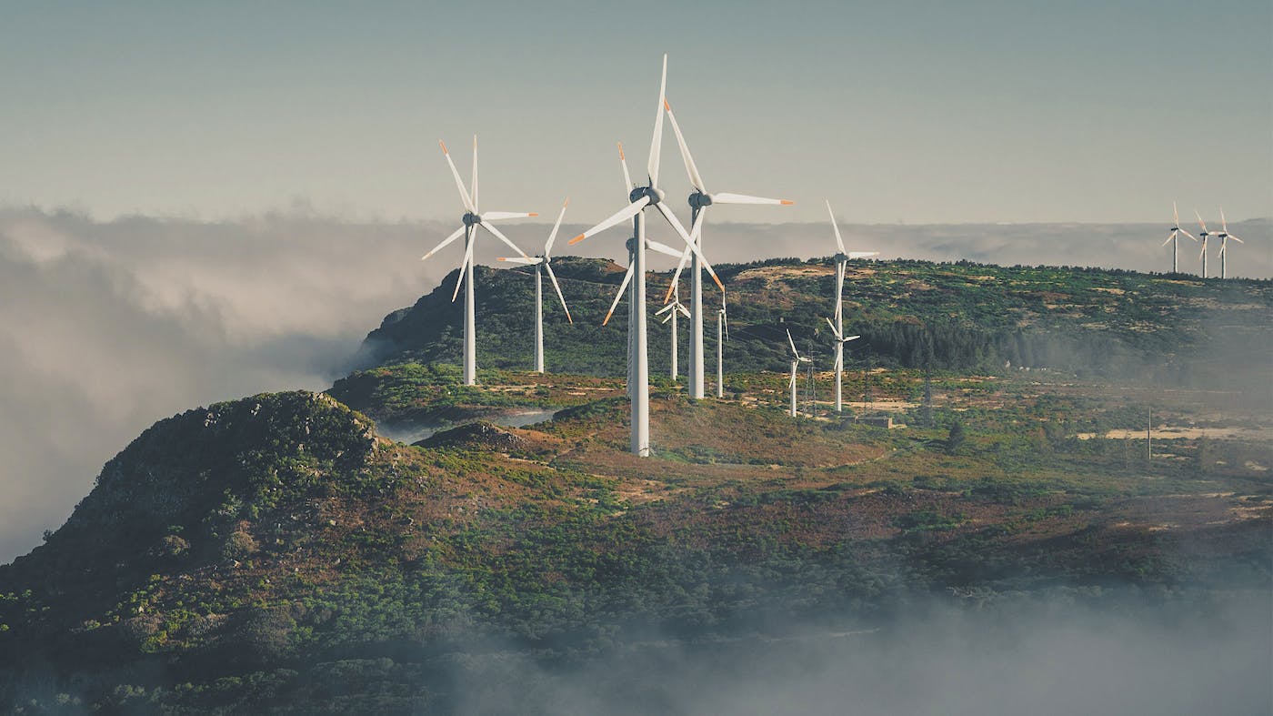 A small wind farm perched on top a lush mountaintop; the mountain is surrounded by pillowy fog.